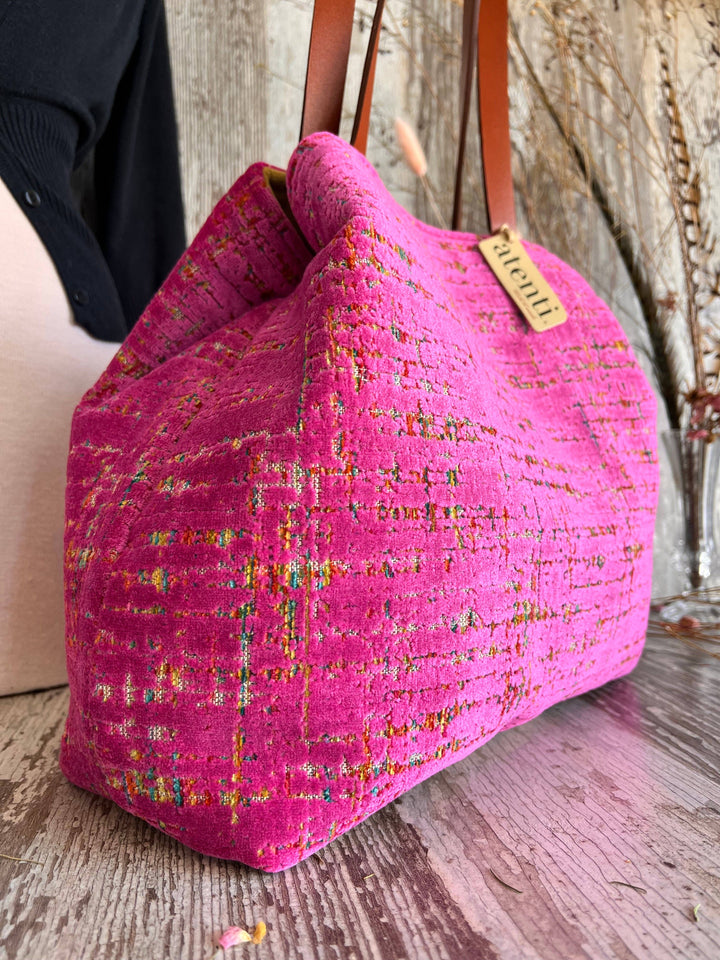 A bright pink tweed handbag with brown leather shoulder straps and a small tan brand tag reading 'PICOLO', shown on a rustic wooden surface.