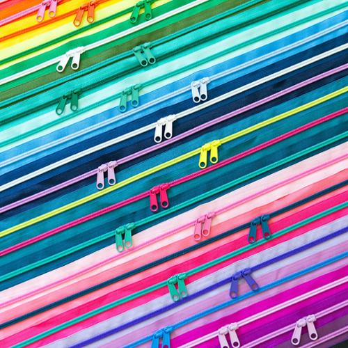 A close-up, diagonal view of multiple colorful zippers arranged in parallel rows, showing a vibrant rainbow spectrum of zipper tape and pull tabs.