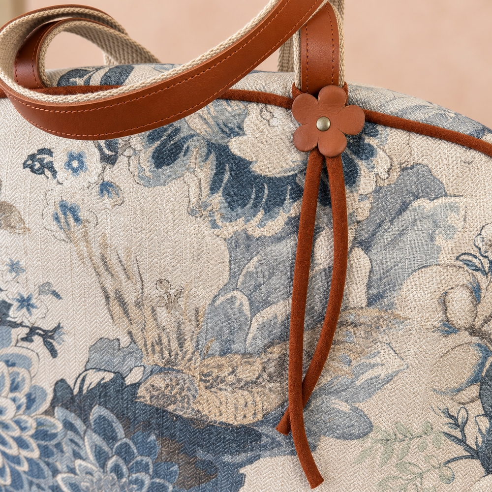 Close-up of a handbag featuring a blue and beige floral tapestry pattern, brown leather trim, and a brown flower-shaped charm with a metal stud.