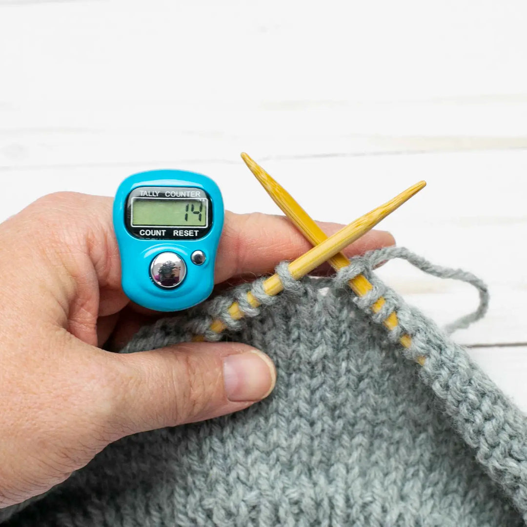 Hand holding a digital knitting counter with yarn and needles on a white background