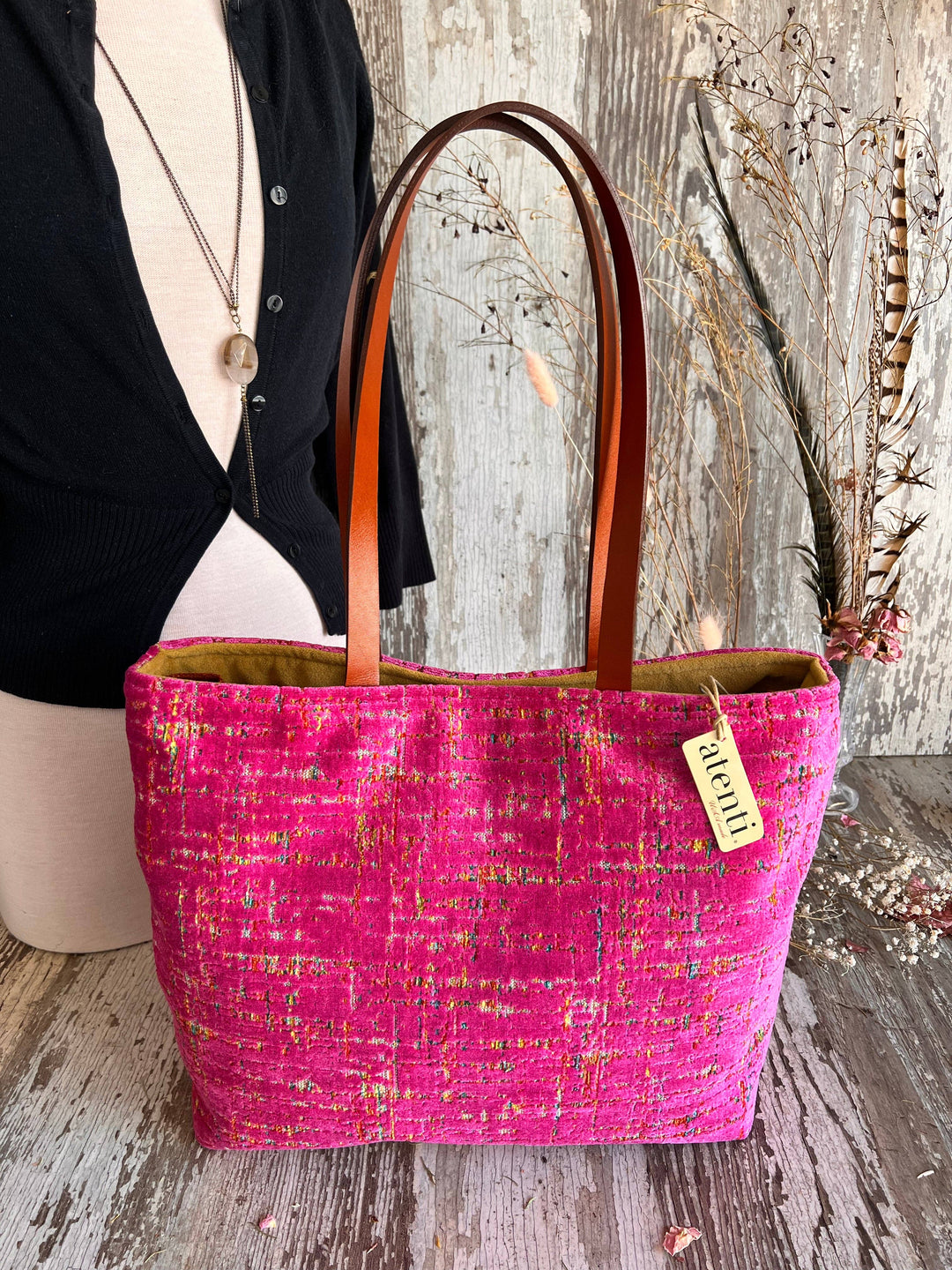 A bright pink woven tote bag with brown leather handles, displayed on a mannequin torso against a rustic wood wall and floor. A small hanging tag reads 'merlot'.