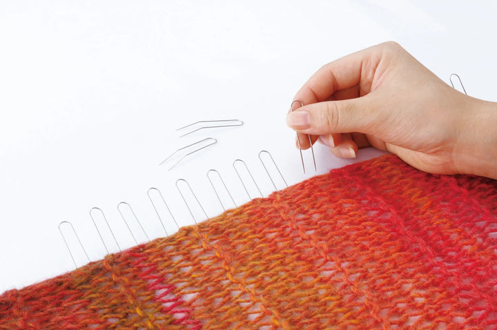 Hand using knitting needles on a red and orange knitted sample against a white background