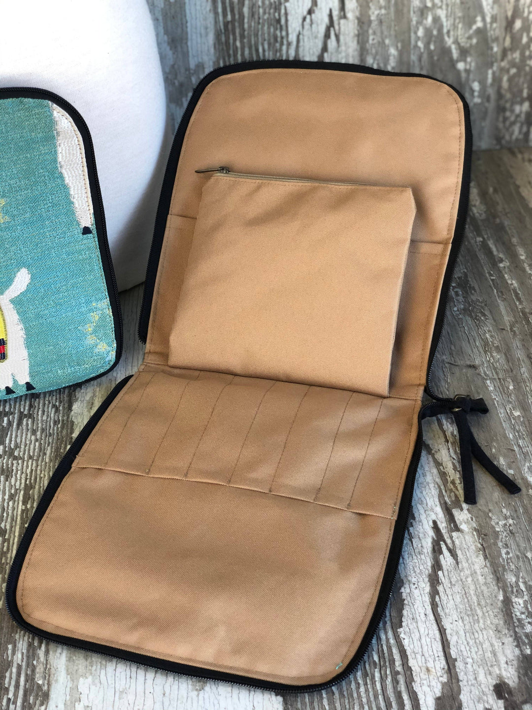 A tan fabric organizer pouch with black trim, shown open on a rustic wood surface. The pouch has a zippered top pocket and multiple stitched compartments.