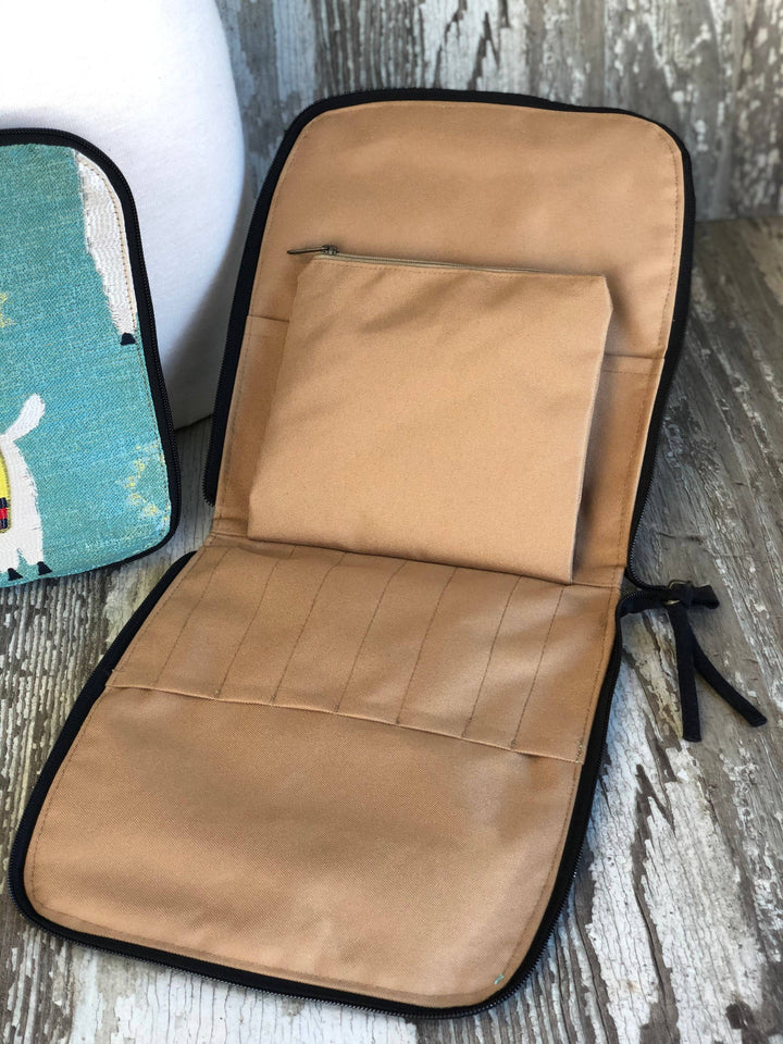 A tan fabric organizer pouch with black trim, shown open on a rustic wood surface. The pouch has a zippered top pocket and multiple stitched compartments.