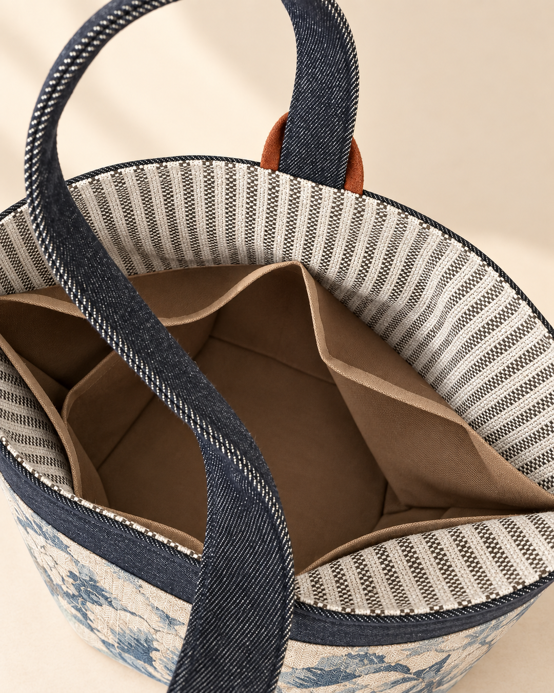 Close-up of a woven tote bag with dark blue denim-like handles, a striped inner lining, and a blue floral pattern on the exterior, shown against a warm beige background.