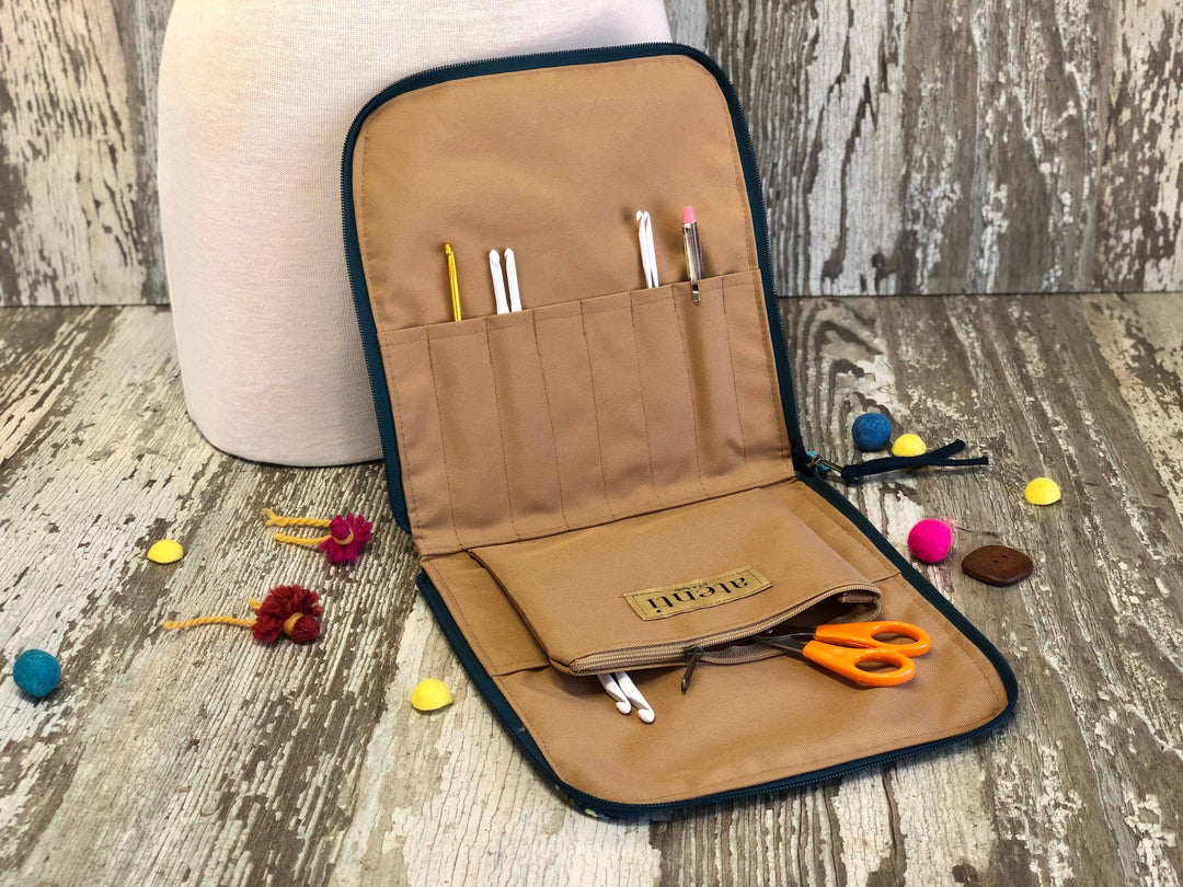 A tan canvas needle case organizer with a dark teal zipper and trim, shown open on a rustic wood surface. The case contains various sewing and crafting tools, including needles, thread, scissors, and a small pouch with a 'SILKTON' brand label.