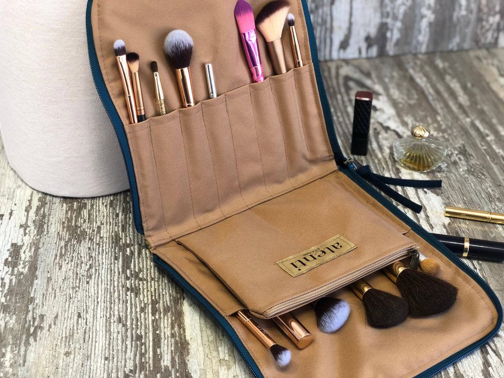 An open tan makeup brush travel case with dark teal trim, holding multiple makeup brushes and a small pouch. The case is placed on a distressed wood surface with a cosmetic bag and other cosmetics nearby.