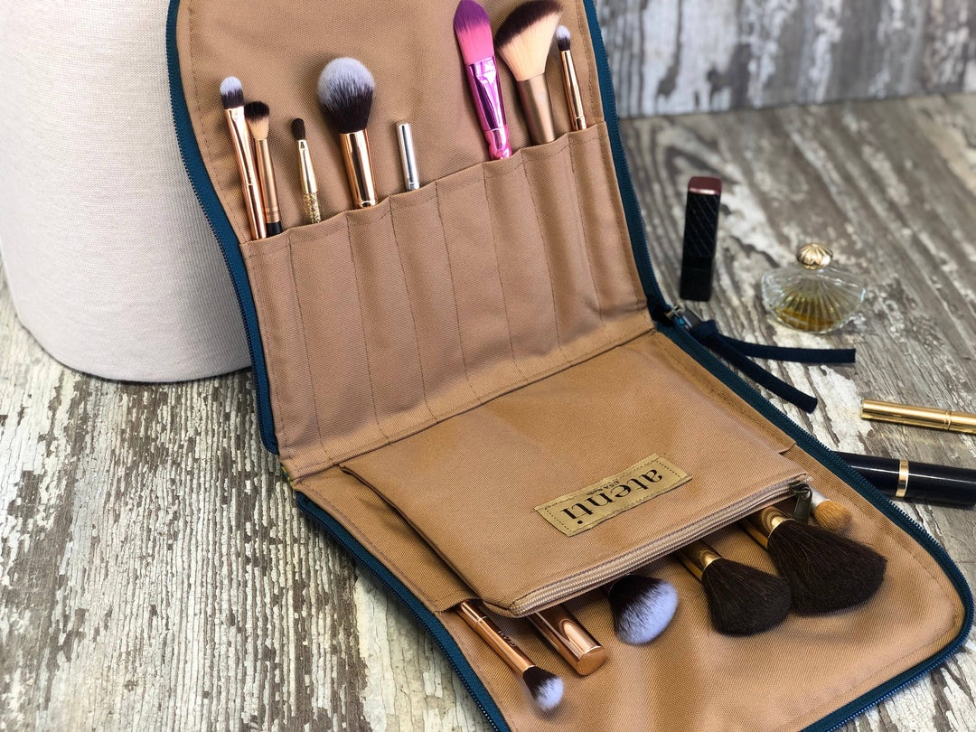 An open tan makeup brush travel case with dark teal trim, holding multiple makeup brushes and a small pouch. The case is placed on a distressed wood surface with a cosmetic bag and other cosmetics nearby.