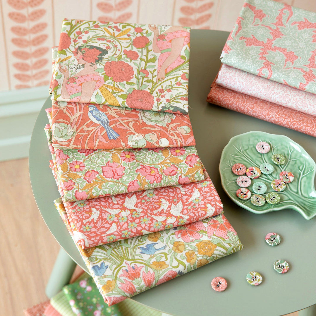 Stacks of colorful fabric on a table with a decorative plate and buttons against a patterned wall.