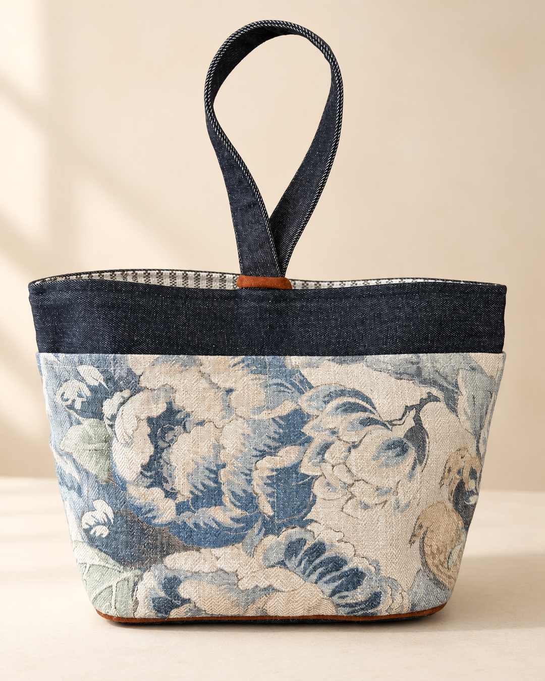 A structured tote bag with a dark blue denim top band and handle, and a lower body featuring a blue and beige floral tapestry pattern, set against a warm beige background.