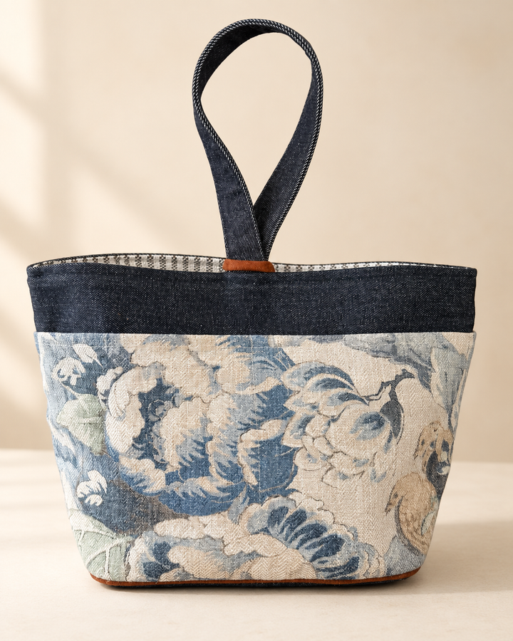 A structured tote bag with a dark blue denim top band and handle, and a lower body featuring a blue and beige floral tapestry pattern, set against a warm beige background.