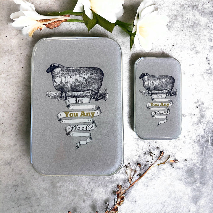 Two metal tins with sheep illustrations and text on a textured surface with flowers.