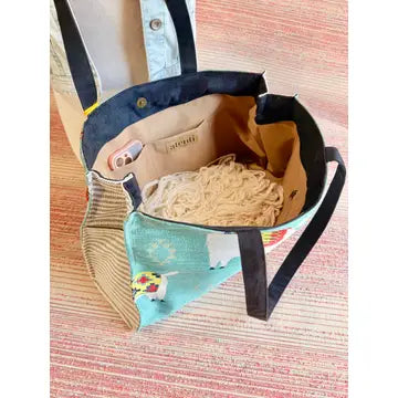 Open tote bag showing beige interior with two pockets - one holding a cell phone and another zippered pouch. Loose bare yarn is in the middle of the bag. 