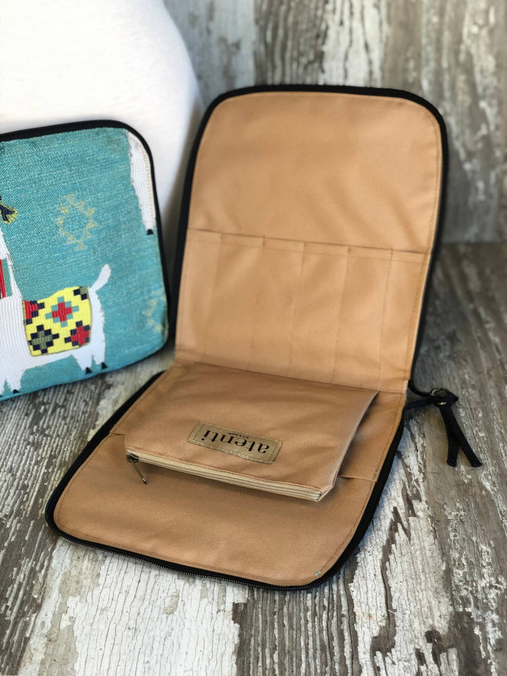 An open tan fabric organizer pouch with black trim, shown on a rustic wood surface. A small notebook or journal with a leather patch reading 'BELIEF' rests inside.