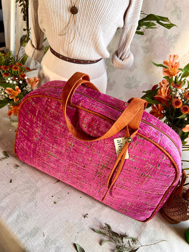 A bright pink textured fabric duffel bag with brown leather handles and trim, displayed on a table with a mannequin torso and floral arrangements in the background.