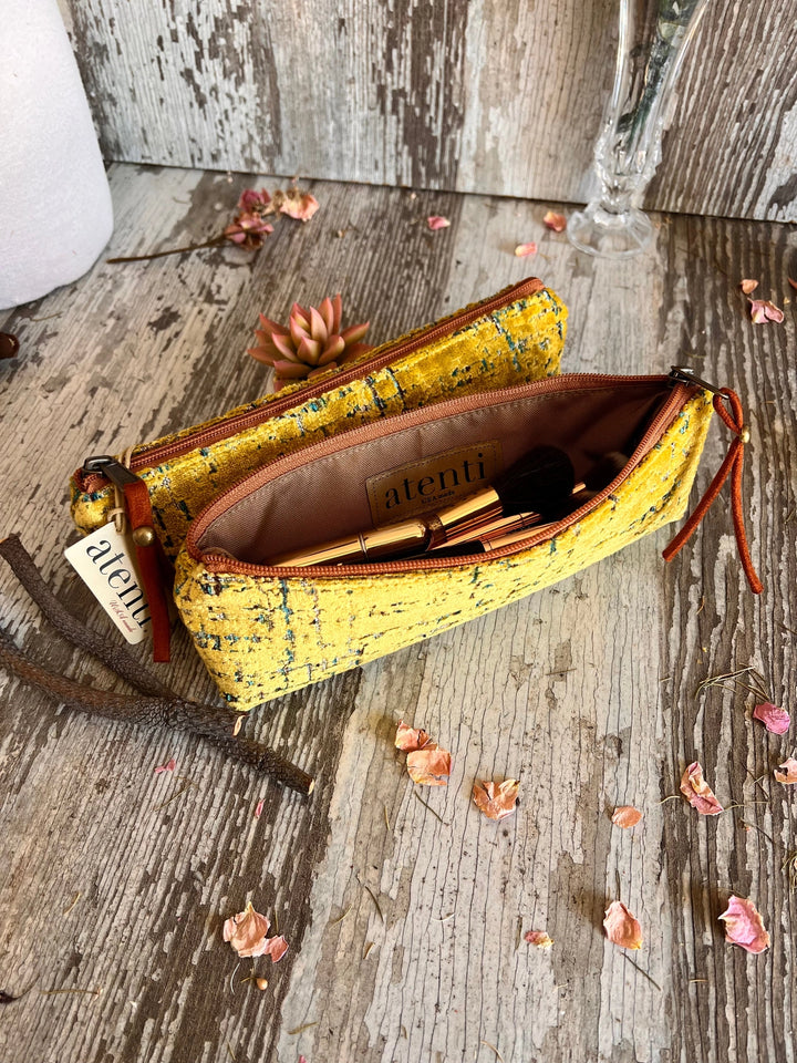 An opened yellow woven fabric zippered pouch with brown leather-like zipper pulls and a small hanging tag, photographed on a rustic weathered wood surface with dried leaves and branches.