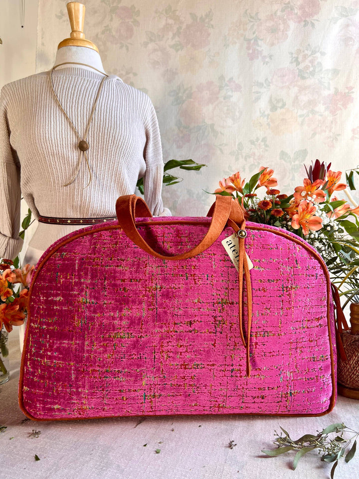 A bright pink textured fabric duffel bag with brown leather handles and a hanging tag, displayed in front of a mannequin wearing a cream ribbed sweater, with orange flowers in the background.