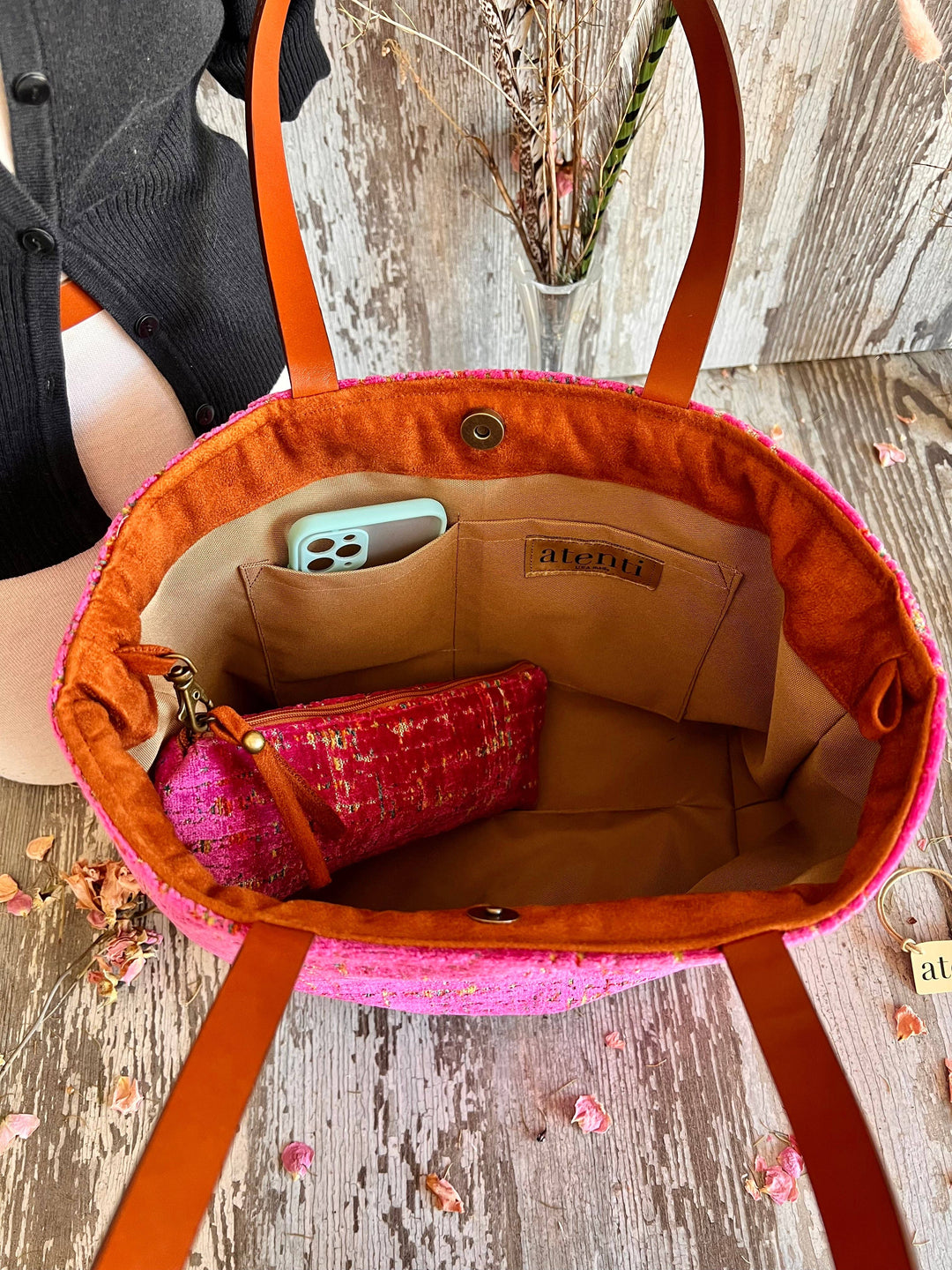 An open woven tote bag with bright pink fabric and orange-brown leather handles, shown from above on a rustic wood surface. Inside the bag is a beige lining, a small pink patterned pouch, and a phone in a light blue case.