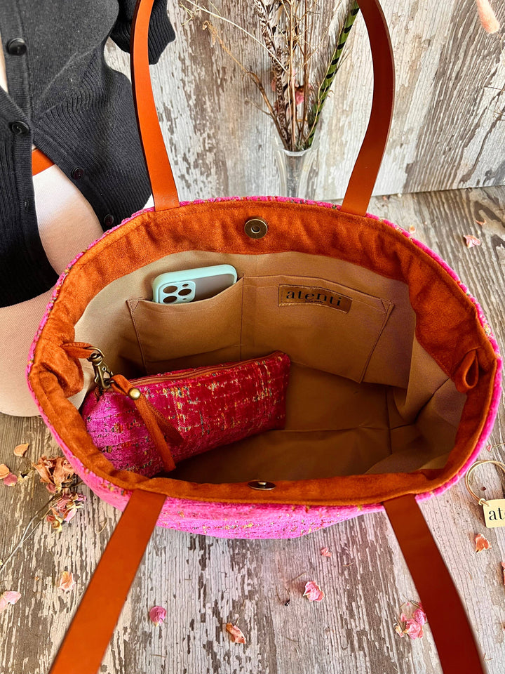 An open woven tote bag with bright pink fabric and orange-brown leather handles, shown from above on a rustic wood surface. Inside the bag is a beige lining, a small pink patterned pouch, and a phone in a light blue case.