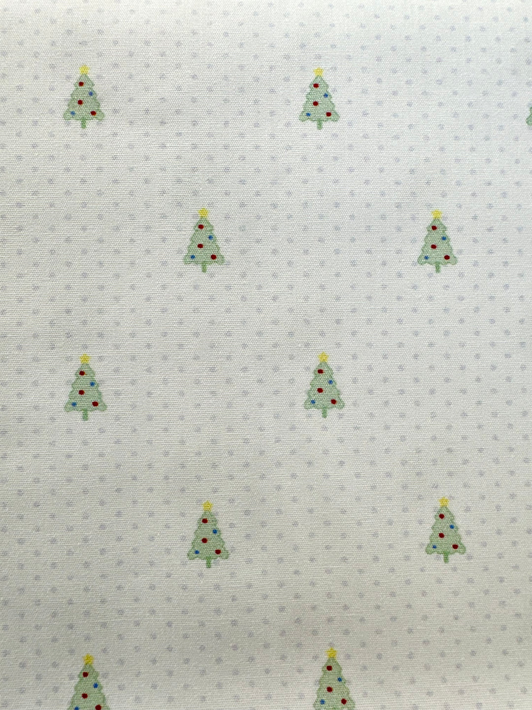 White fabric with a repeating pattern of small green Christmas trees decorated with red ornaments and yellow stars, interspersed with tiny blue polka dots.