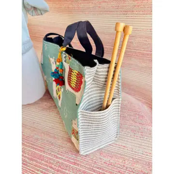 Blue tote bag with llamas on the front, showing a gray and white striped side panel with a pouch and two knitting needles sticking out of it. 