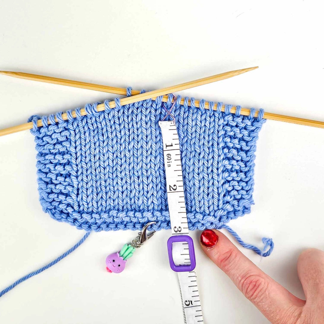 a blue swatch of knitting is active on wooden knitting needles with the Work Until... Measuring tape stitch marker on the needles and the lock guide in the 3 inch spot.