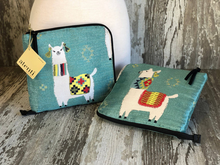 Two teal zippered pouches with embroidered llama designs, displayed on a rustic wood surface with a white ceramic vase behind them. One pouch has a yellow 'atenti' tag attached.