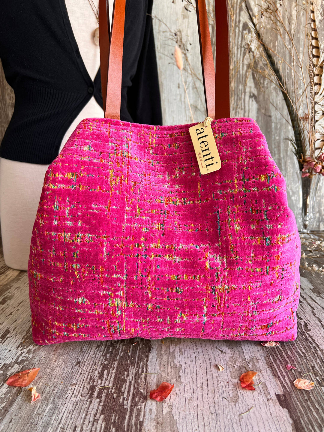A bright pink tweed shoulder bag with brown leather straps, displayed on a rustic wooden surface with a mannequin torso in the background. A small paper tag on the bag reads 'alchemist'.