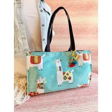 Blue tote bag with black handles and a colorful tassel displaying llamas wearing a colorful blanket. 