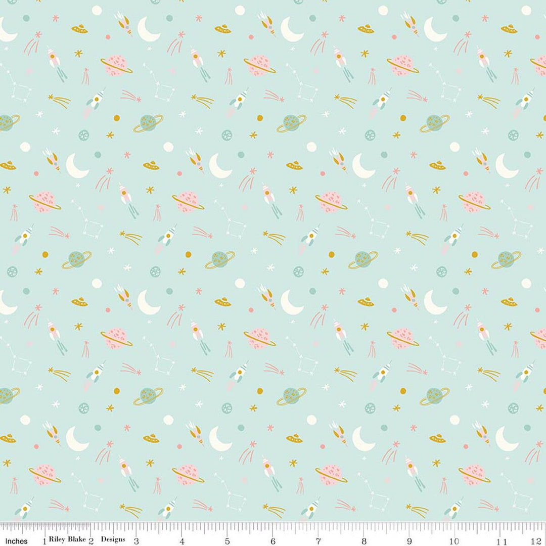 A repeating fabric pattern on a pale mint green background featuring whimsical space motifs including pink and white rockets, gold and pink UFOs, white crescent moons, small stars, and tiny planets. A ruler along the bottom edge shows the scale of the print.