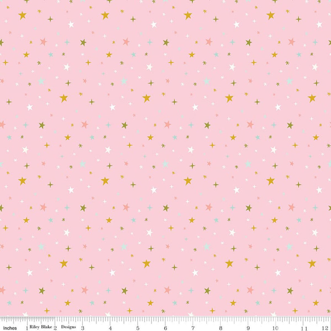 Pink fabric with a repeating pattern of small stars and plus signs in white, gold, light blue, and peach, shown with a ruler along the bottom edge.