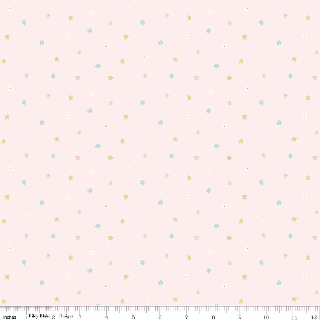 A repeating fabric pattern featuring scattered blossoms in pink, green, blue and white  on a light pink background, shown with a ruler along the bottom edge indicating scale.