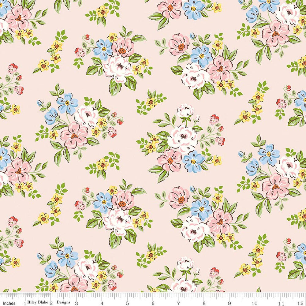 A repeating floral pattern on a pale blush pink background, featuring clusters of white, pink, and light blue flowers with green leaves and small yellow blossoms. A white fabric ruler is visible along the bottom edge.