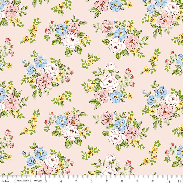 A repeating floral pattern on a pale blush pink background, featuring clusters of white, pink, and light blue flowers with green leaves and small yellow blossoms. A white fabric ruler is visible along the bottom edge.