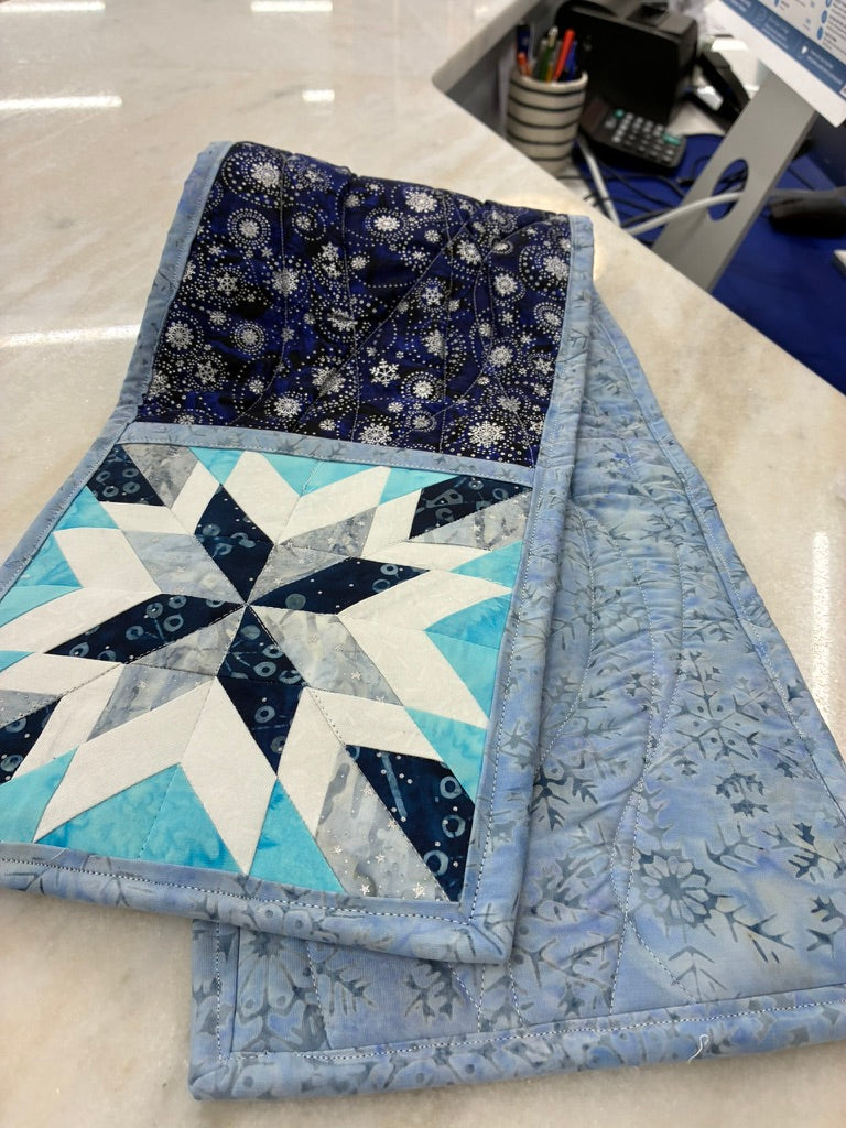 A folded quilted textile with a starburst patchwork design in light blue, white, and navy, paired with a dark navy patterned section and a light blue floral fabric side, photographed on a light countertop.