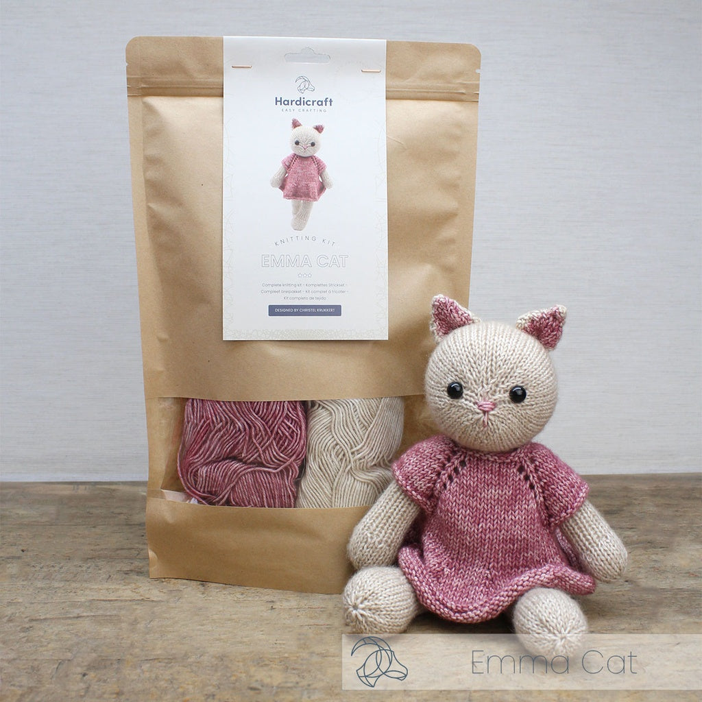 Knitted cat toy with pink dress next to a package of yarn on a wooden surface.