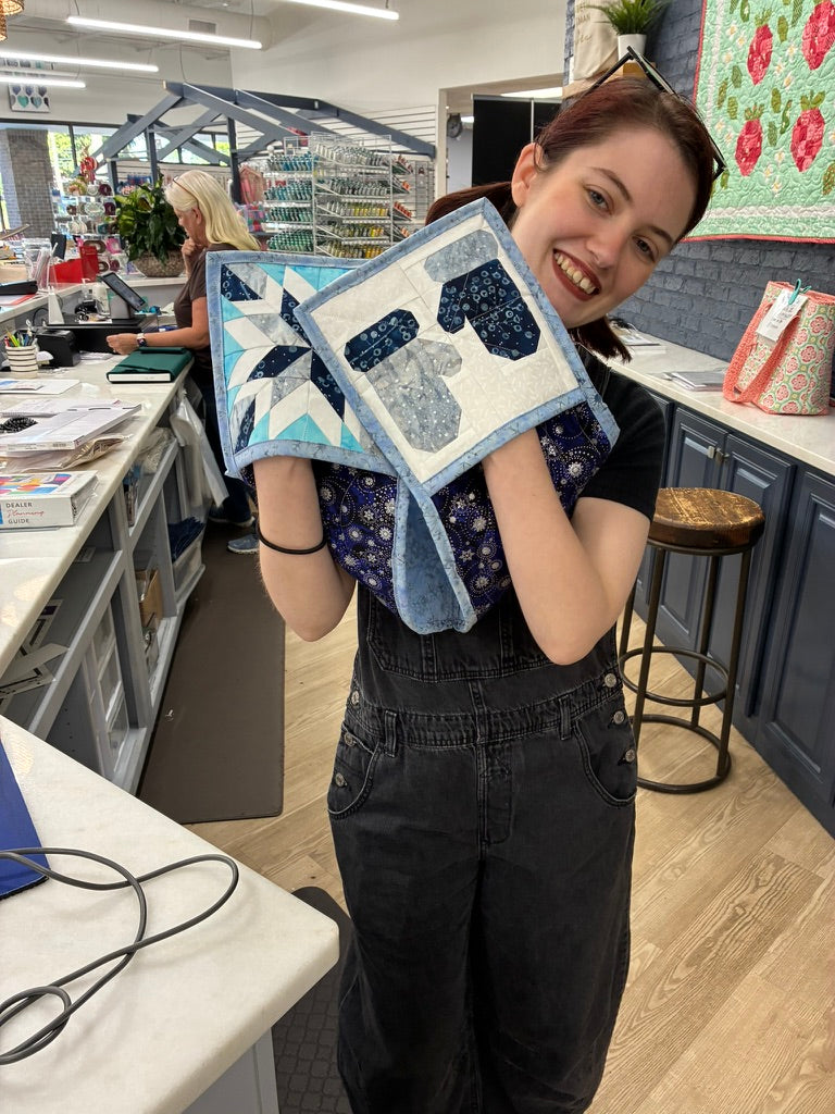 A smiling woman in a sewing or quilting studio holds up two small quilted fabric blocks. The blocks feature blue and white patterns, including a mitten design on one piece and a starburst design on the other.