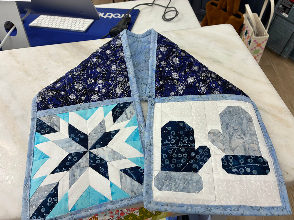 A handmade quilted potholder laid flat on a white marble tabletop. The garment features a blue and white starburst quilt on one side and a pair of mittens on the other, with a blue patterned yoke at the top.