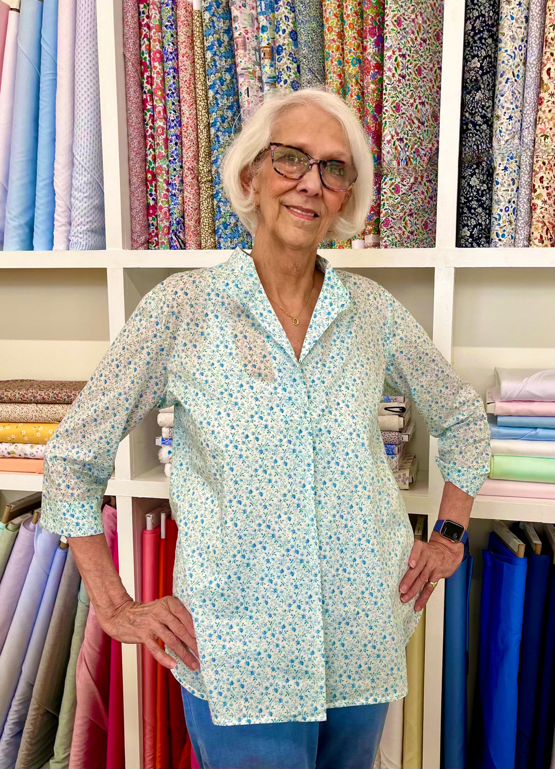 An older woman standing in front of fabric swatches on a wall, wearing a floral print peasant blouse.