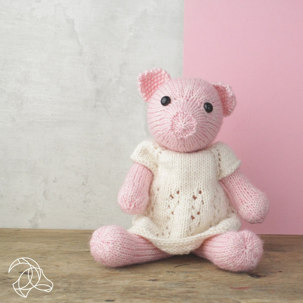 Knitted pink bear toy with a white dress on a wooden surface against a light gray and pink background.