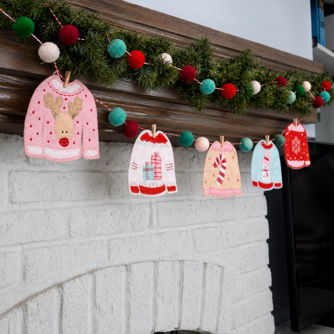 Mahogany fireplace mantel hanging green garland, multi colored ball garland and hanging from that are small embroidered sweaters. 