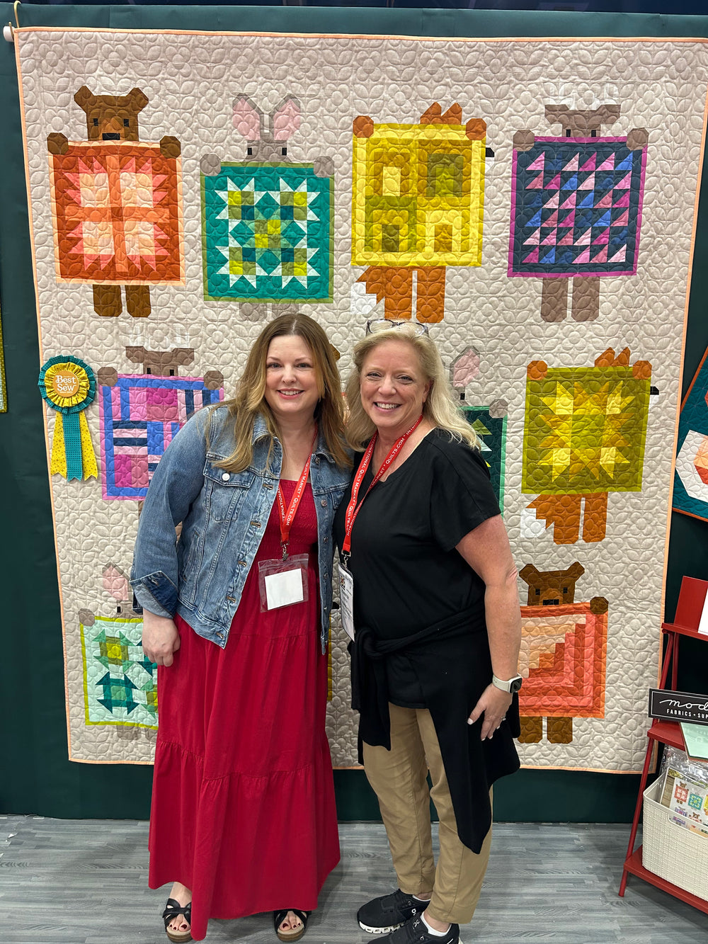 Two women standing in front of a colorful quilt with animal designs.