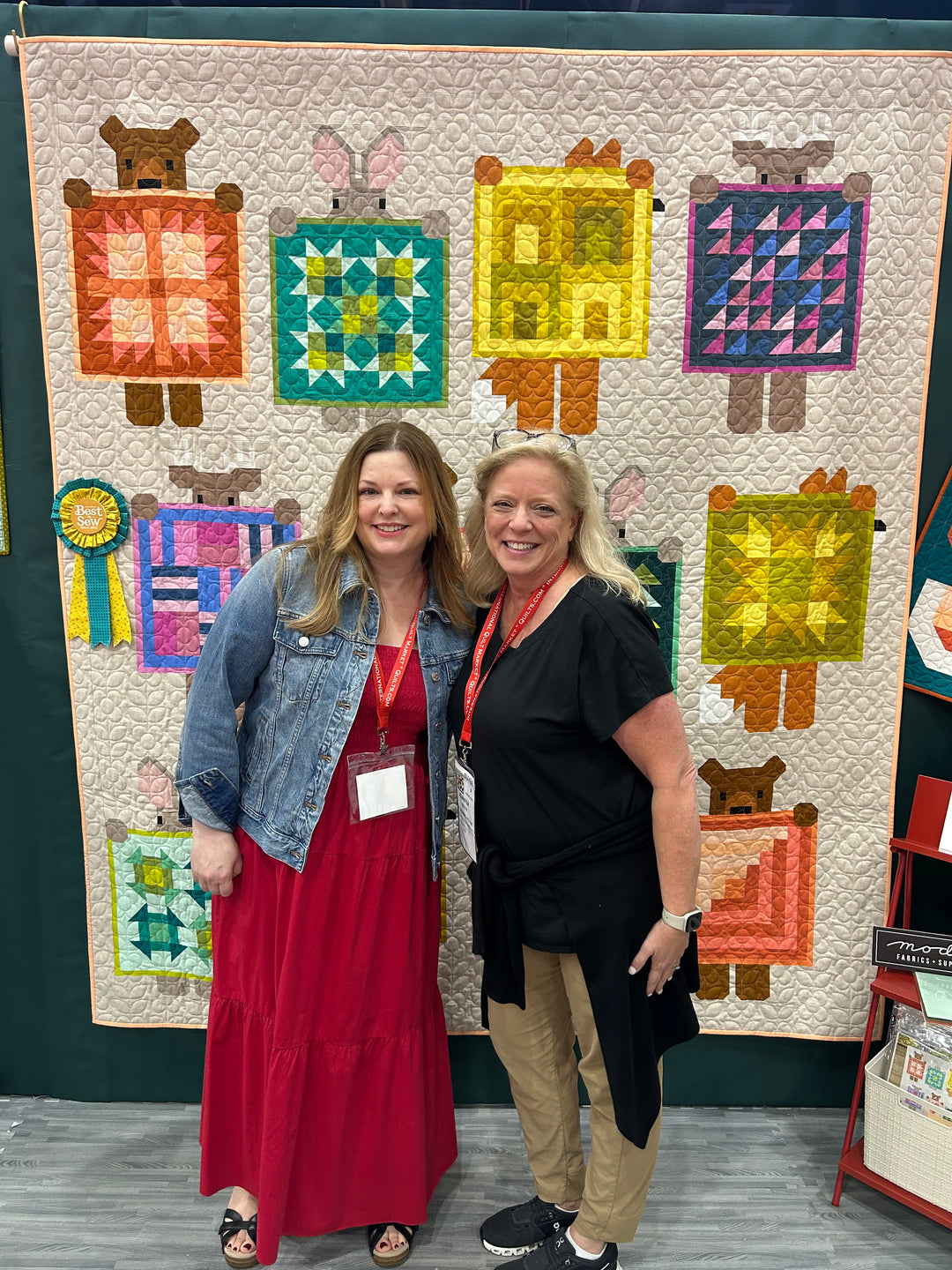 Two women standing in front of a colorful quilt with animal designs.