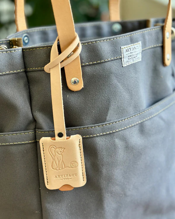 Gray tote bag with a tan leather strap and tag featuring a logo and text.