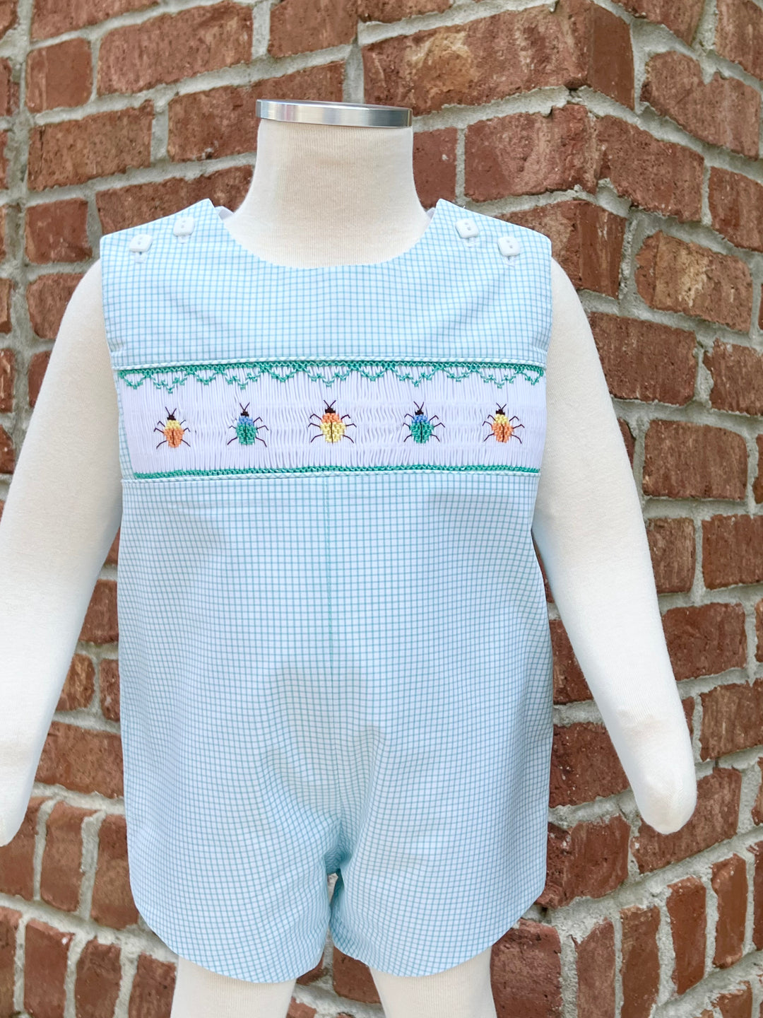 Light blue checkered romper with embroidered designs on a mannequin against a brick wall.