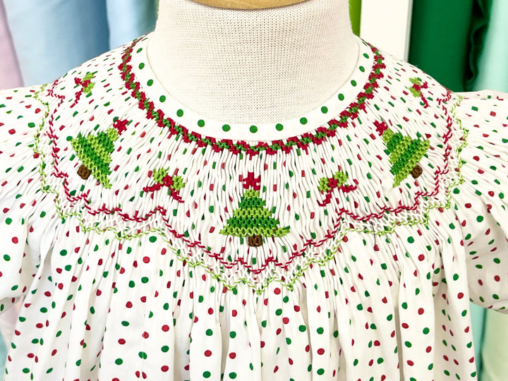 A close-up of a bishop dress on a mannequin smocked with green Christmas trees and bows on red and green polka dot fabric.