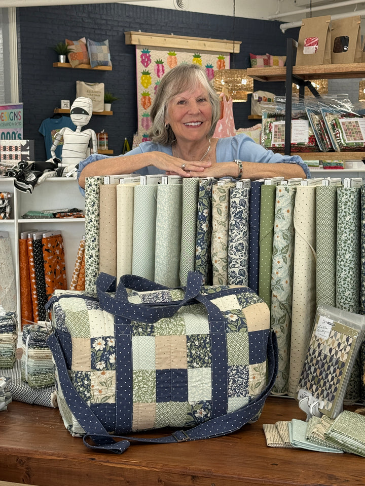 A woman smiling behind a display of fabric rolls and a large patchwork tote bag in navy and cream tones, set inside a craft or fabric store.