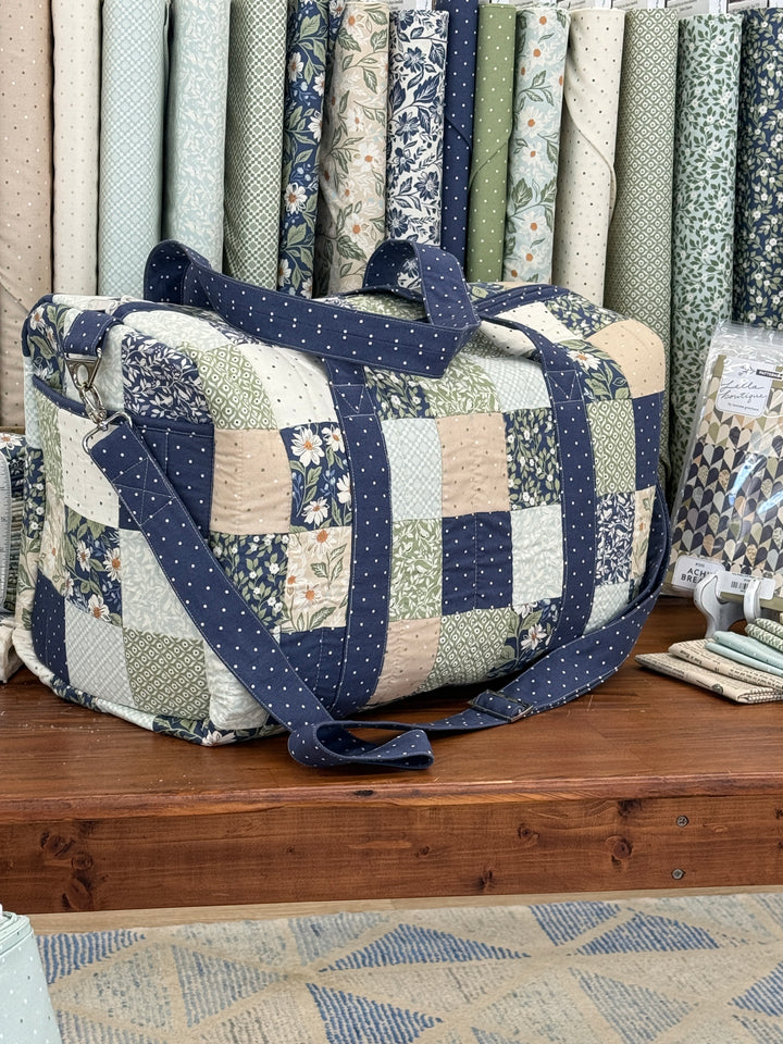 A handmade patchwork quilted fabric tote bag with navy blue polka dot straps, displayed on a wooden shelf next to stacks of folded quilts.