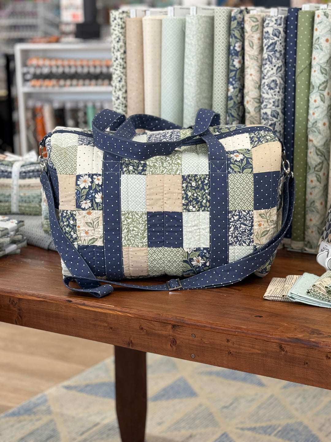 A patchwork quilted fabric duffel bag with navy blue straps and a multicolor patchwork body, displayed on a wooden table in a craft shop with fabric rolls in the background.