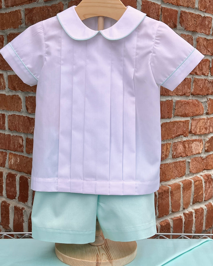 Children's outfit with white shirt and light blue shorts on a wooden hanger against a brick wall.